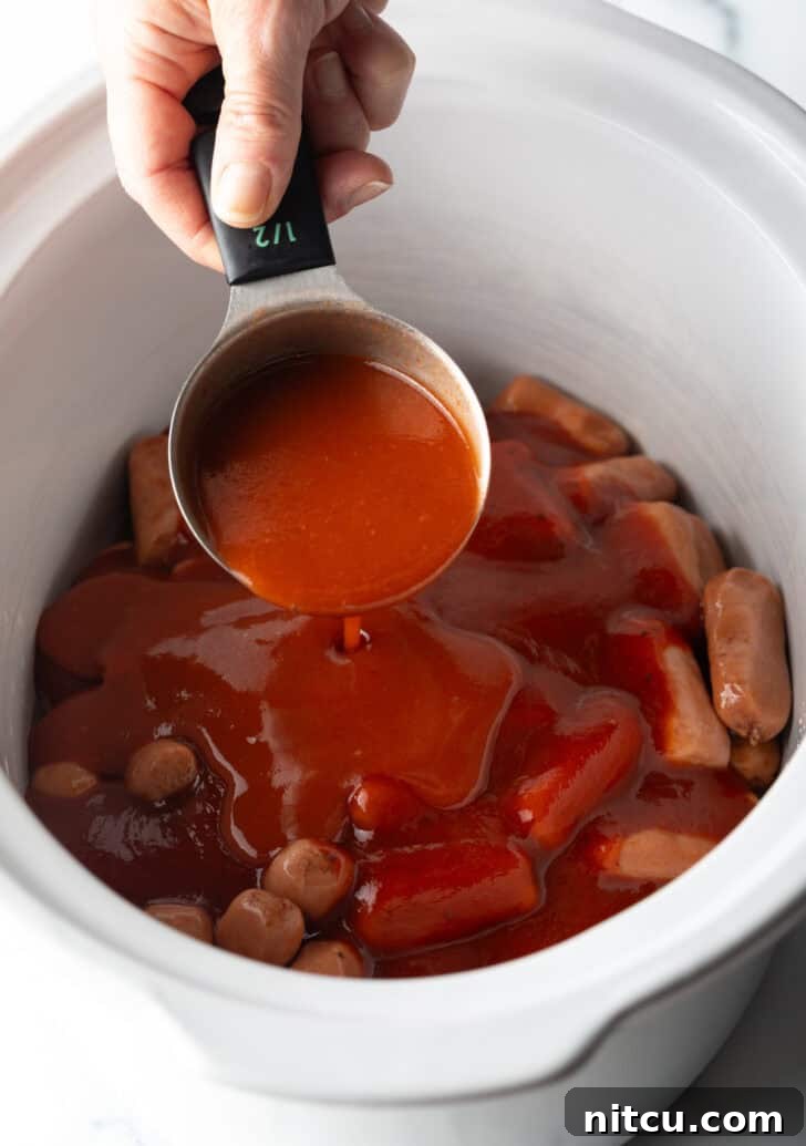 Hand pouring hot sauce from a measuring cup into a slow cooker with smokies.