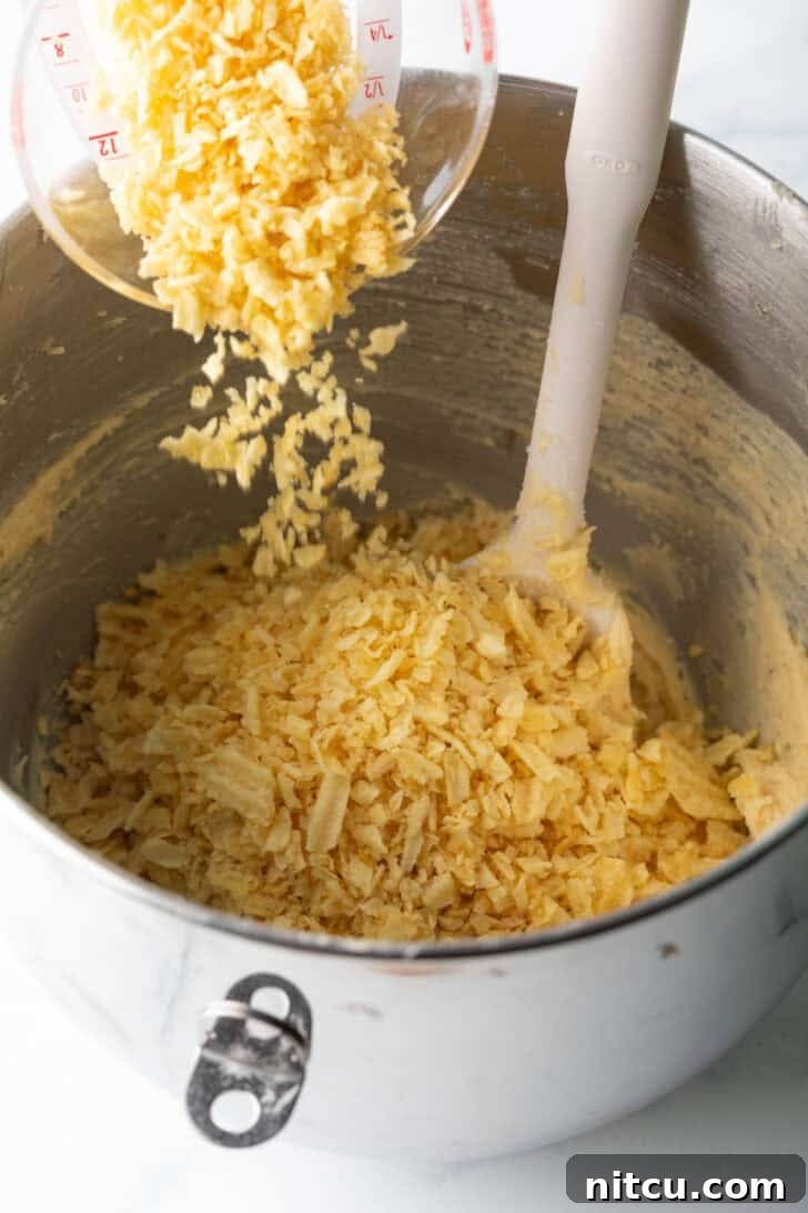 Crushed potato chips being gently poured over chocolate chip cookie dough in a mixing bowl.