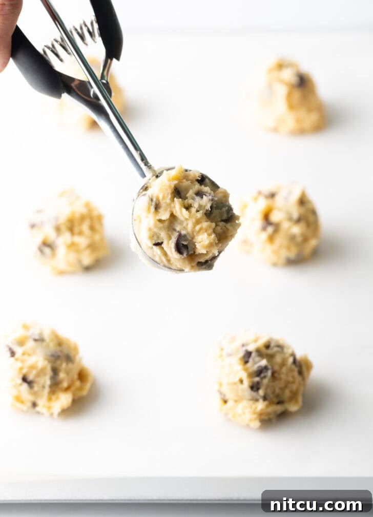 Cookie dough scooped into a metal cookie scoop, with several rounded cookie dough balls arranged on a baking sheet below.