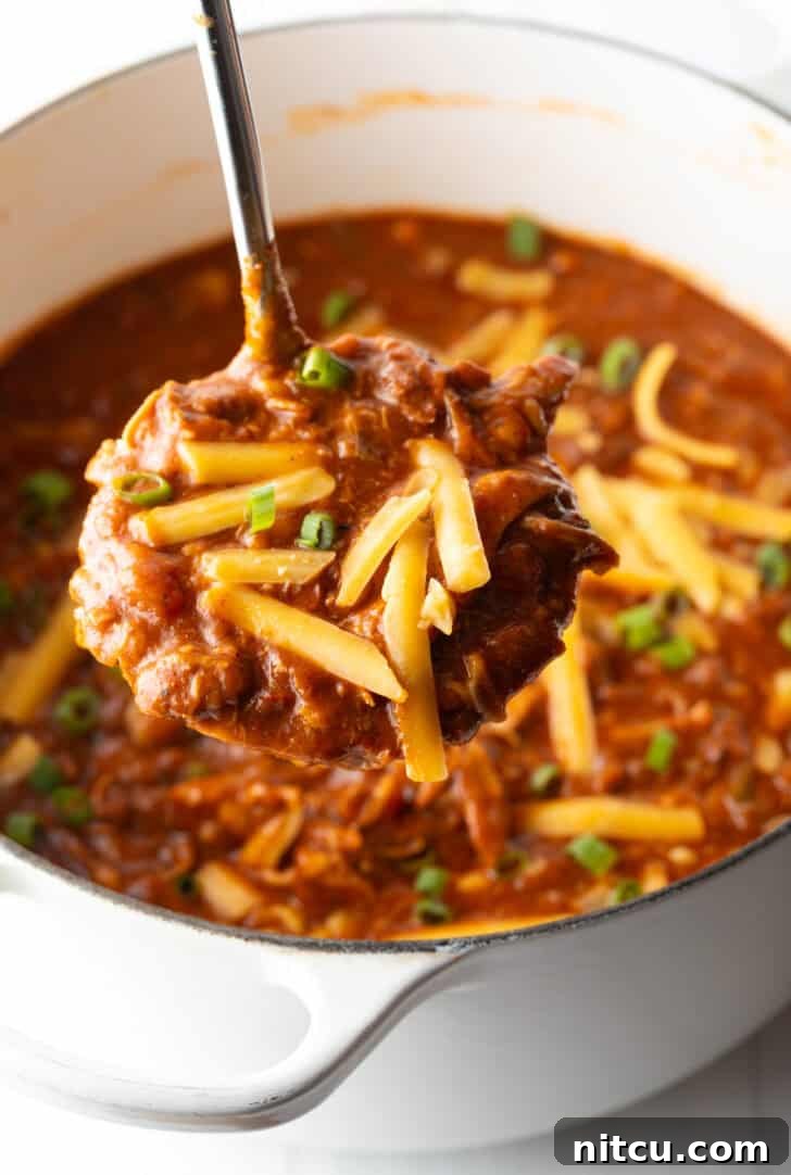 A ladleful of rich chicken chili, generously topped with shredded cheddar cheese, held up to the camera with steam gently rising, highlighting its warmth and appetizing texture.