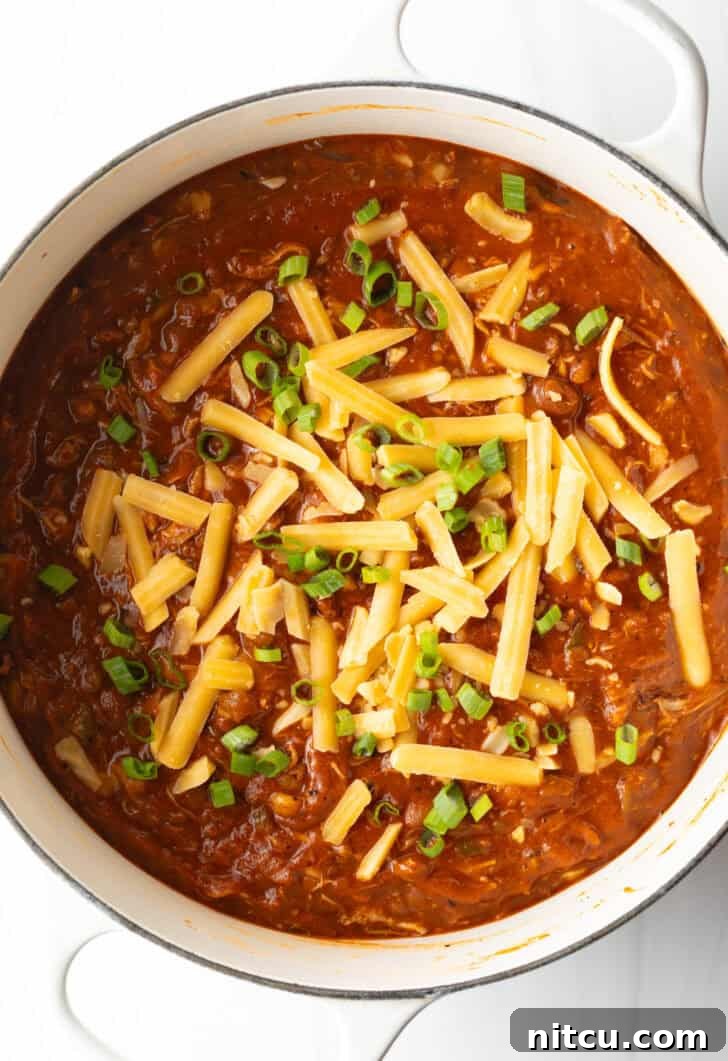 A pristine white soup pot, seen from directly above, brimming with the inviting chicken chili, exquisitely garnished with finely chopped green onions and a generous layer of shredded cheddar cheese, presenting a visually appealing and comforting meal.