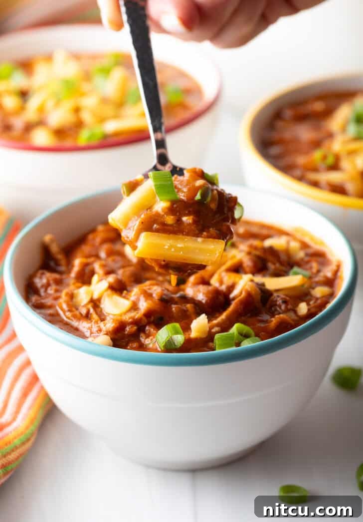 A close-up view of a spoon digging into a white bowl filled with creamy shredded chicken, bean, and salsa chili, revealing the hearty texture and rich blend of ingredients within.