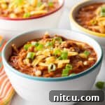 A bowlful of rich chicken chili, beautifully topped with fresh chopped green onion and a generous portion of shredded cheddar cheese, ready to be enjoyed.