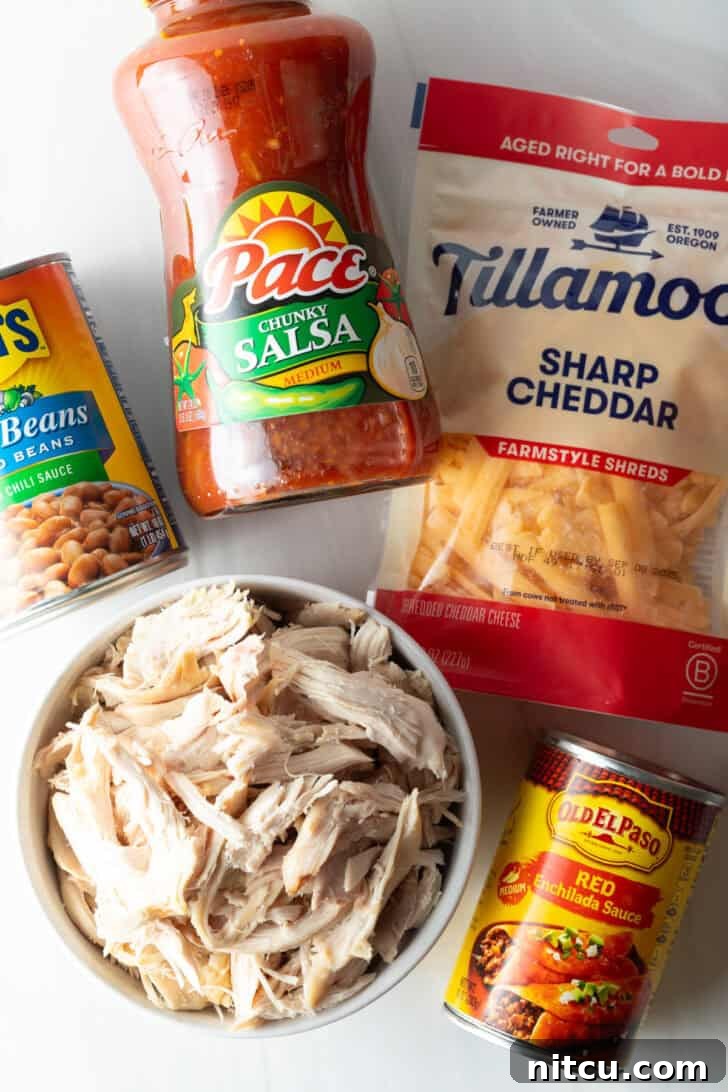 A top-down view displaying all five core ingredients for the chicken chili recipe: a bowl of finely shredded cooked chicken, a bottle of chunky salsa, a large can of red enchilada sauce, two cans of pinto beans in sauce, and a bag of premium shredded cheddar cheese.