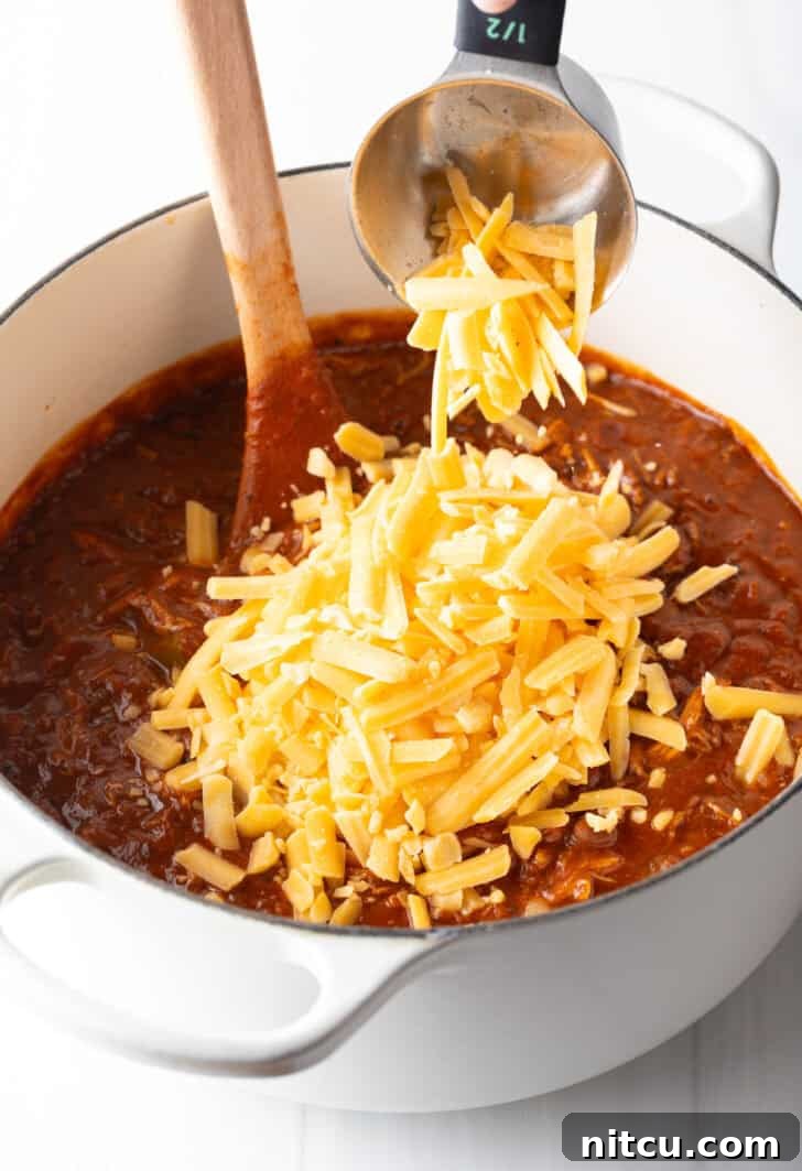 A generous amount of shredded cheddar cheese being carefully added to the large pot of warm chili, poised to melt and create a creamy, luxurious texture that defines this easy chicken chili.