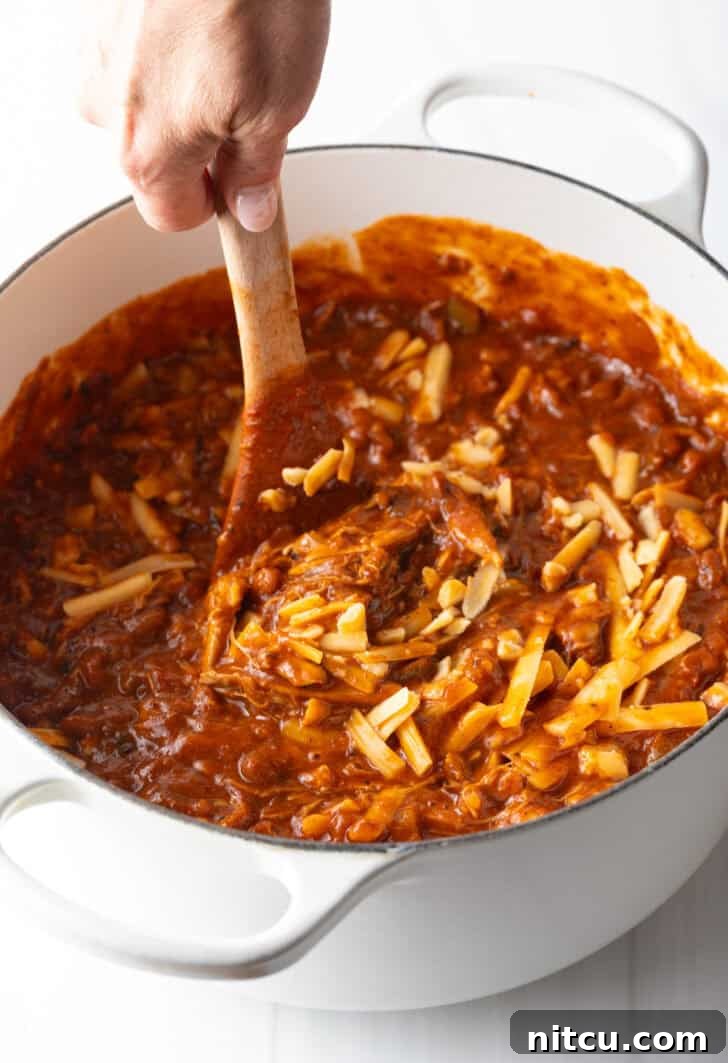 A sturdy wooden spoon gently stirring the shredded cheese into the hot chili within the pot, demonstrating the crucial step of incorporating the cheese off the heat for a perfectly smooth and creamy finish.