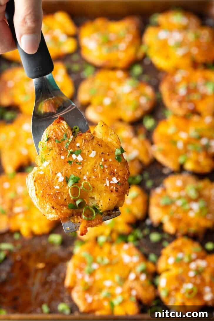 Top down view of 15 golden smashed potatoes on a baking sheet. A hand with a spatula is holding a crispy smashed potato to camera.