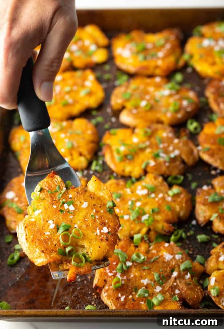 Top down view of 15 golden smashed potatoes on a baking sheet. A hand with a spatula is holding a crispy smashed potato to camera.