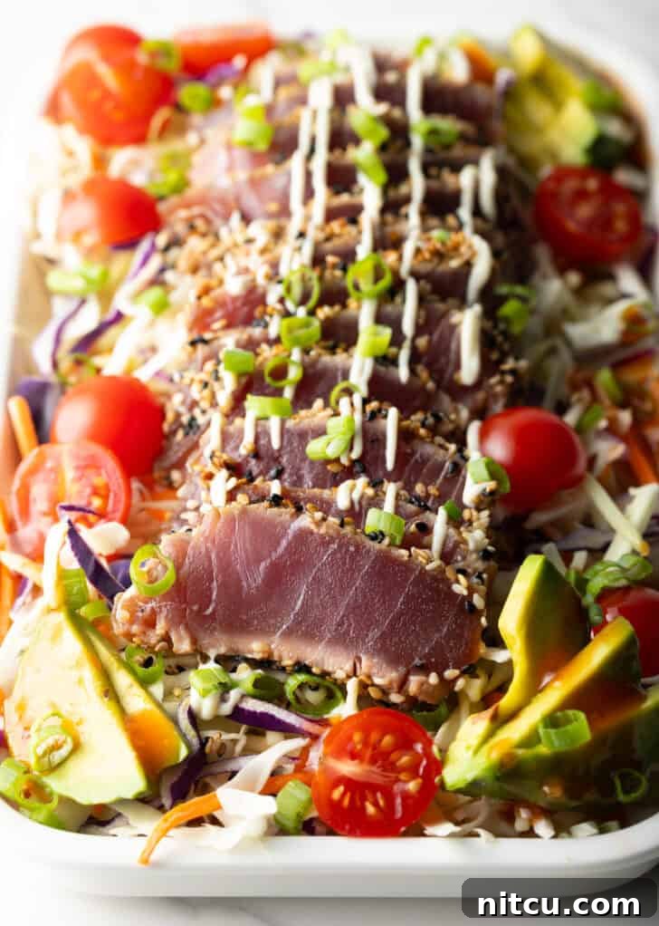 Large white rectangular dish loaded with fresh salad greens, halved cherry tomatoes, avocado slices, delicate slices of seared ahi tuna, chopped green onions, and a drizzle of dressing.