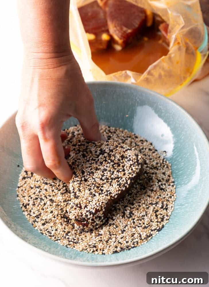 A hand gently pressing a raw piece of ahi tuna steak into a bowl filled with tricolor sesame seeds for an even coating.