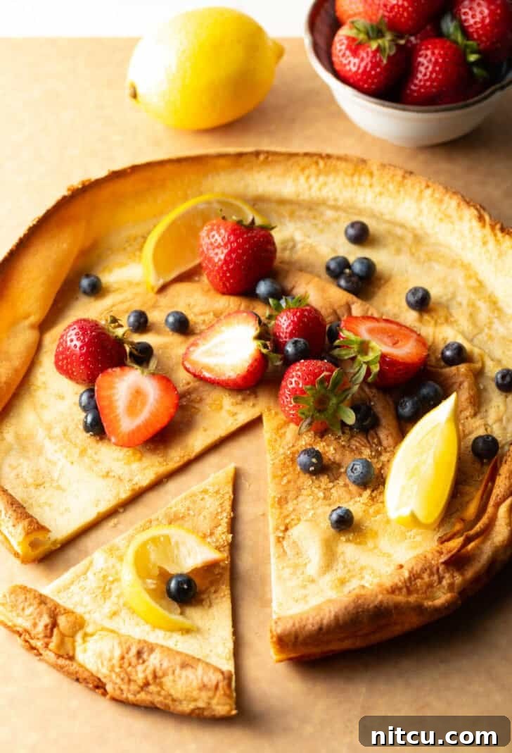 A baked Dutch Baby pancake on a wooden cutting board, adorned with fresh strawberries and blueberries. One wedge has been pulled aside, showing its tender layers.