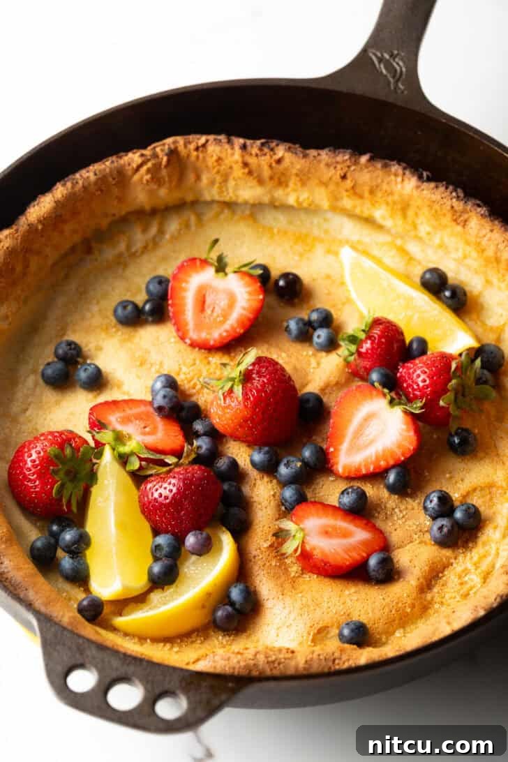A full Dutch Baby pancake in a cast iron skillet, garnished with an abundance of fresh strawberries, blueberries, and delicate lemon slices.