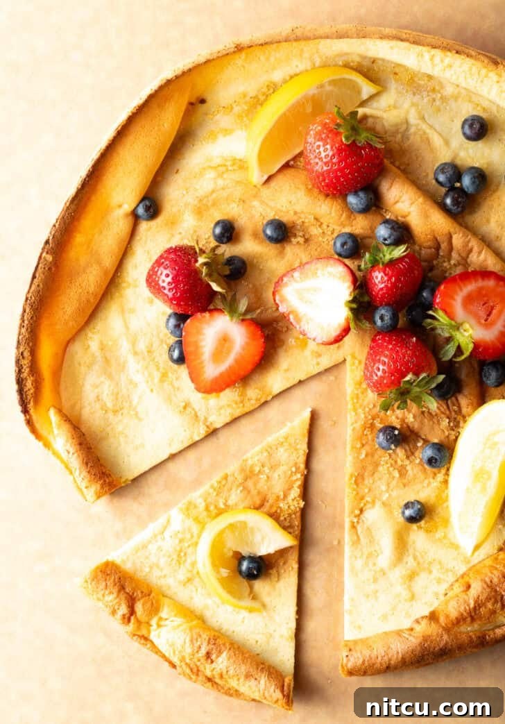 Top-down view of a Dutch Baby on a cutting board, garnished with fresh strawberries and blueberries. A slice has been removed, revealing its inviting texture.