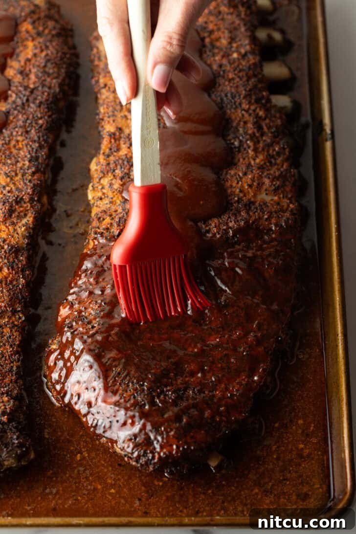 St. Louis-style ribs on an oven tray being brushed with barbecue sauce for the final glaze.