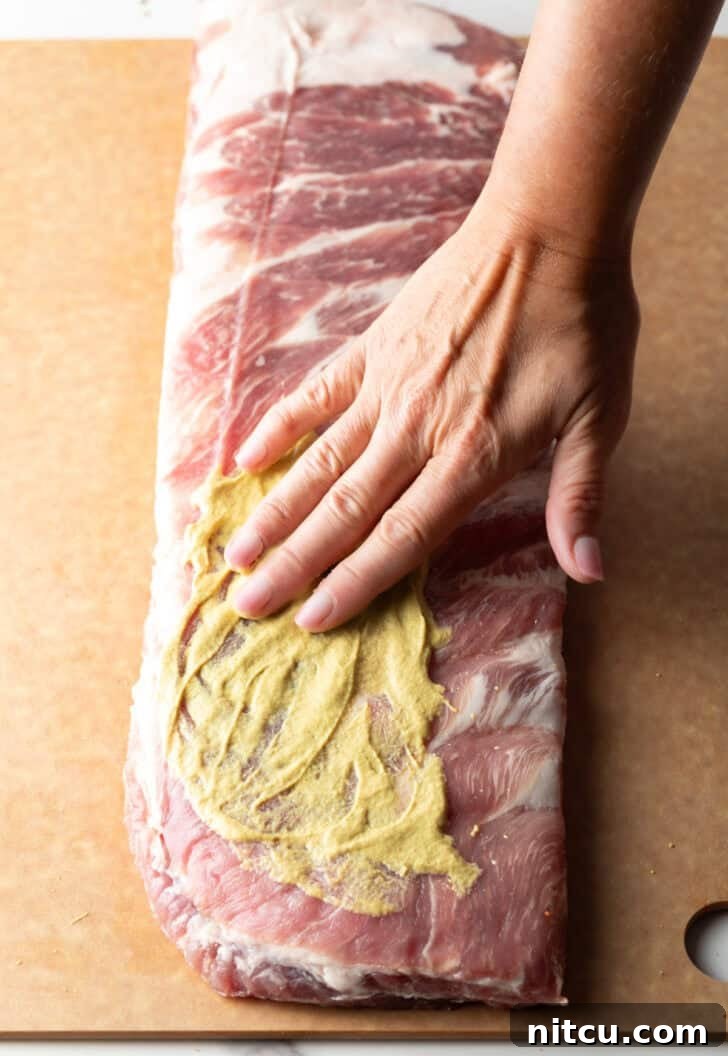 Overhead shot of a raw rack of St. Louis pork ribs being generously smeared with Dijon mustard.