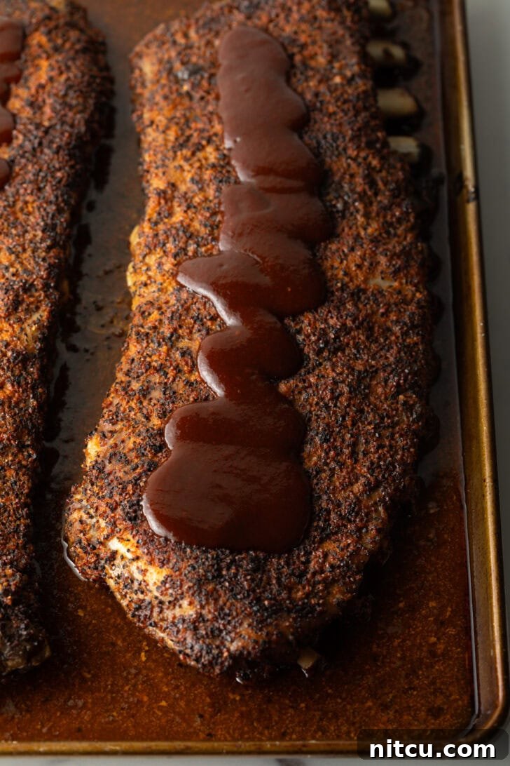 Cooked ribs on an oven tray being drizzled with barbecue sauce.