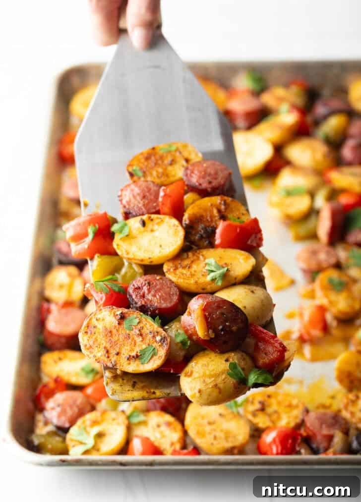 Metal spatula holding up a scoop of roasted kielbasa, potatoes, and red peppers from the sheet pan.
