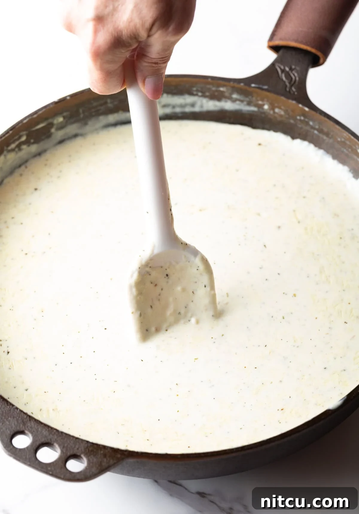 A hand stirring creamy Alfredo sauce in a large stainless steel skillet with a white spatula.