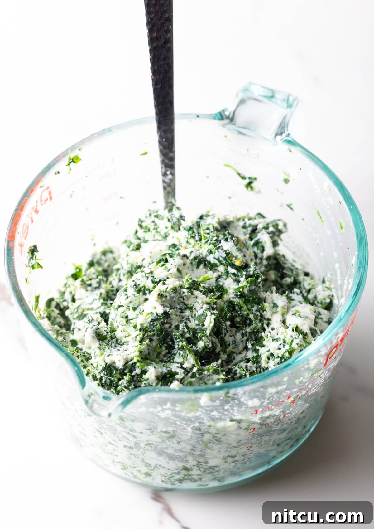 A close-up of thawed frozen spinach and creamy ricotta cheese combined in a glass measuring cup, ready for the lasagna filling.