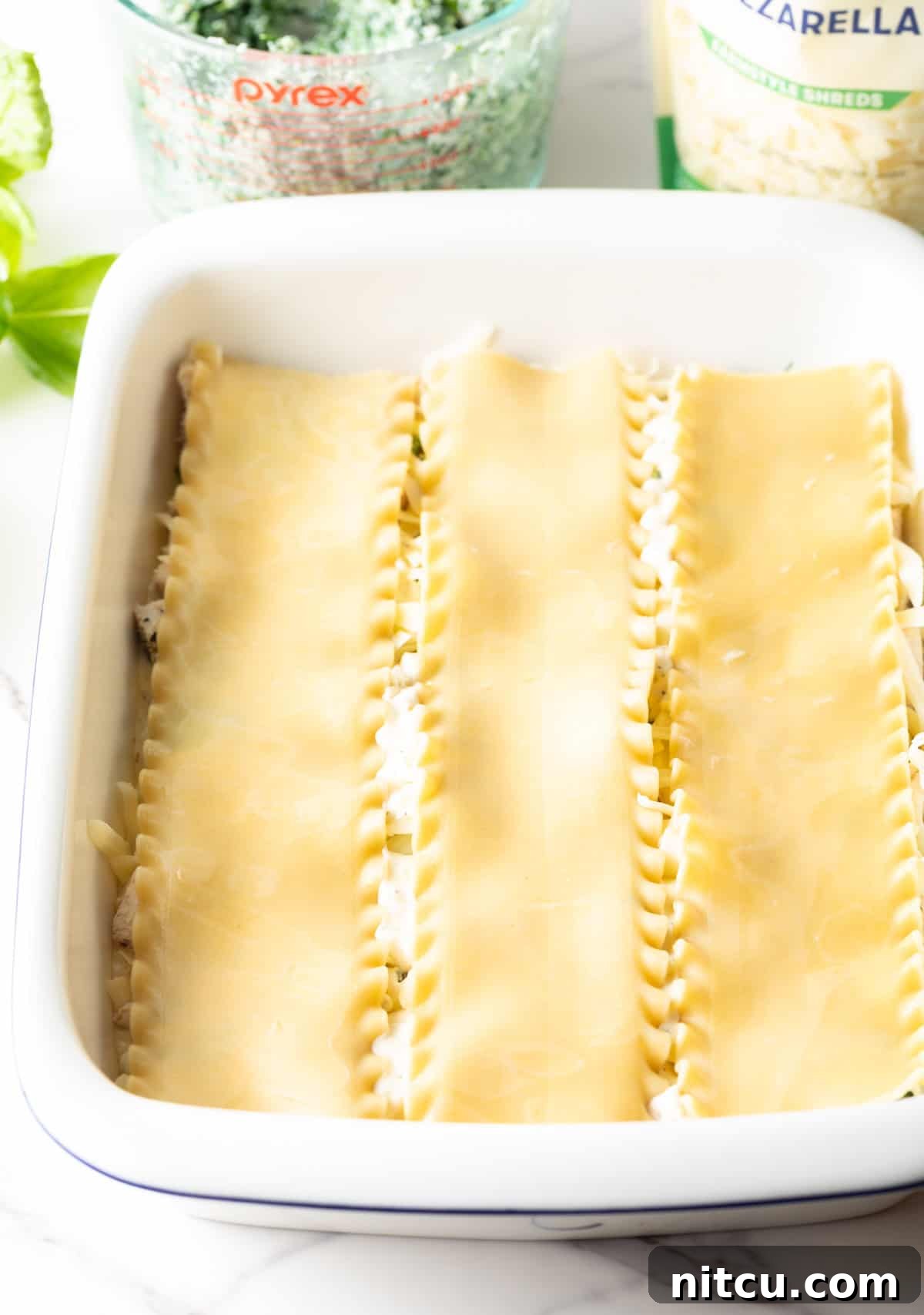 A top-down view of lasagna noodles being carefully placed in a white rectangular casserole dish as the base layer.