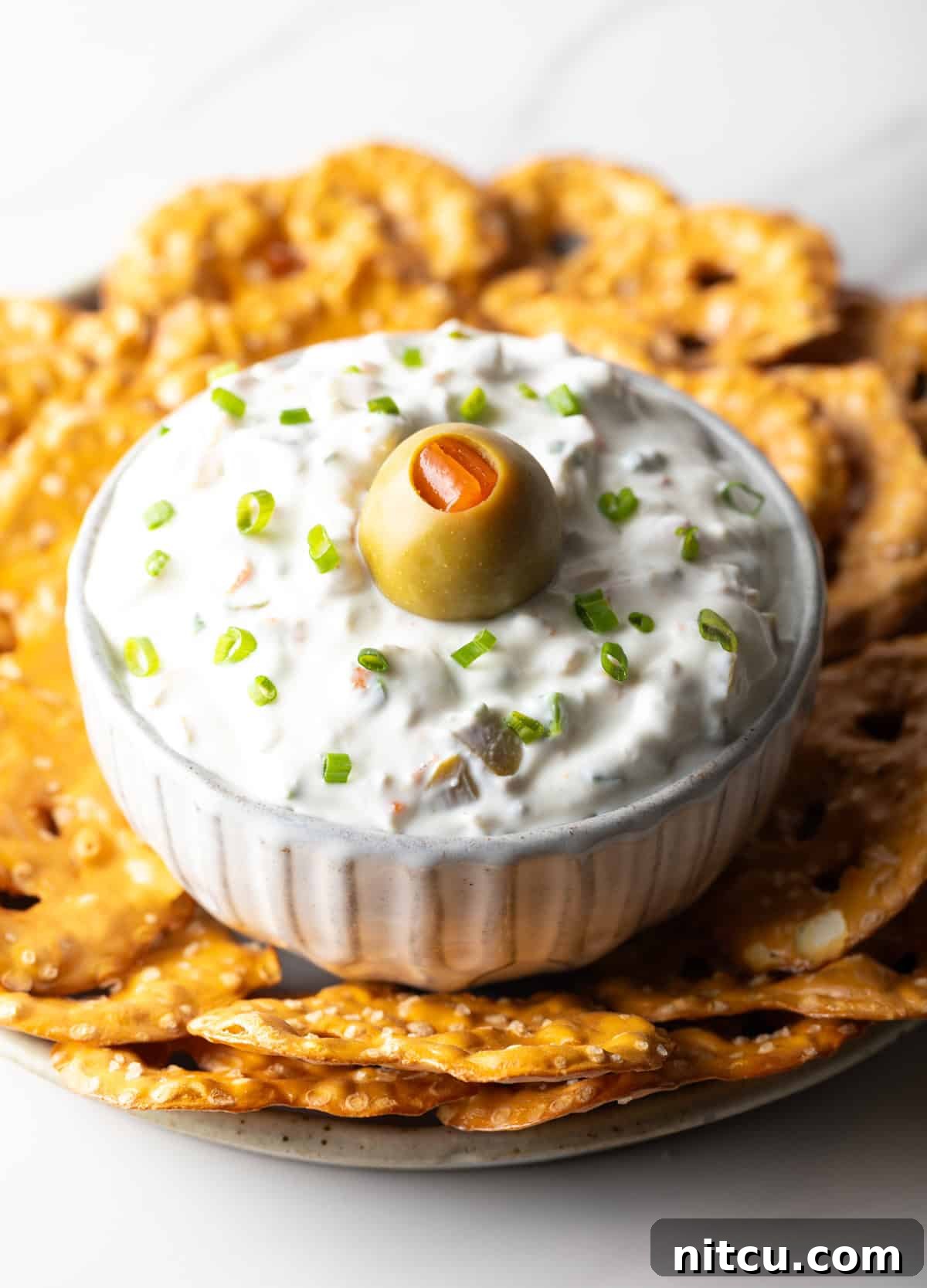 White bowl of dirty martini dip topped with one green olive, surrounded by pretzel chips, perfect for parties.