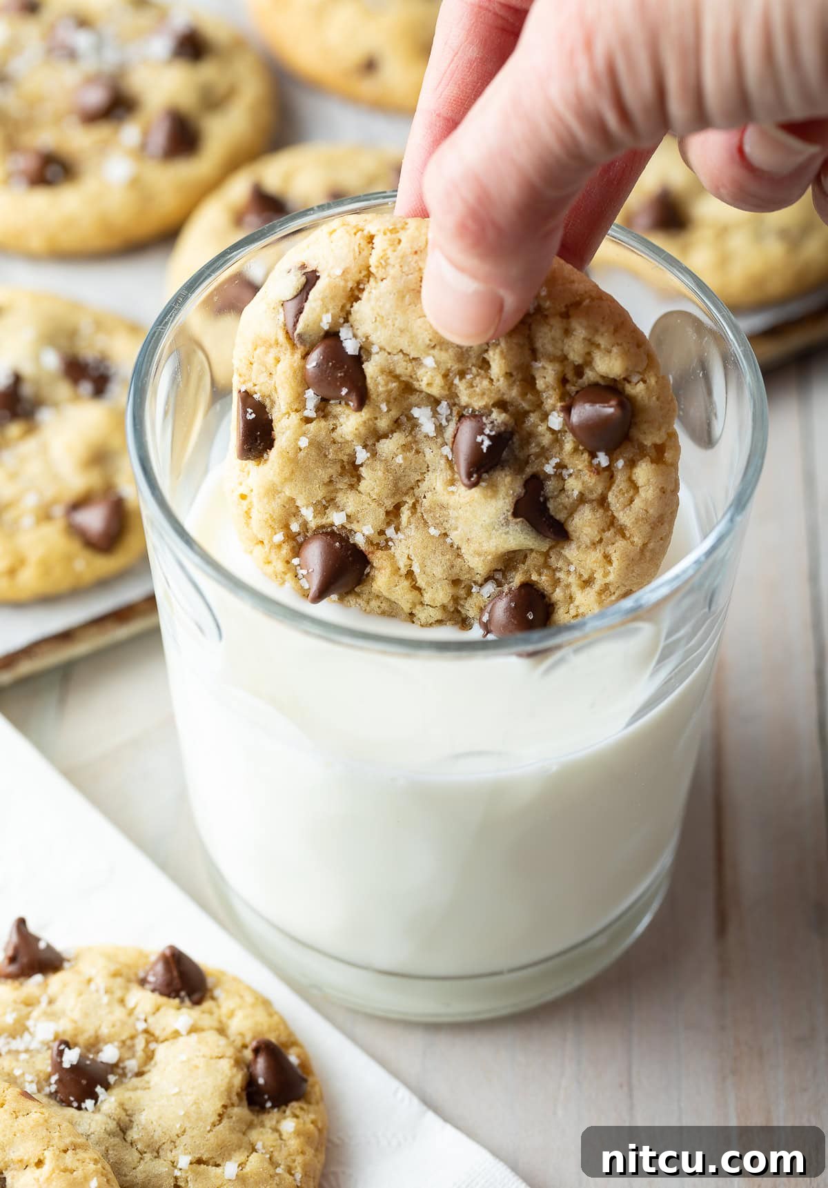 A hand dunking a freshly baked salted chocolate chip cookie into a refreshing glass of milk, a classic pairing.
