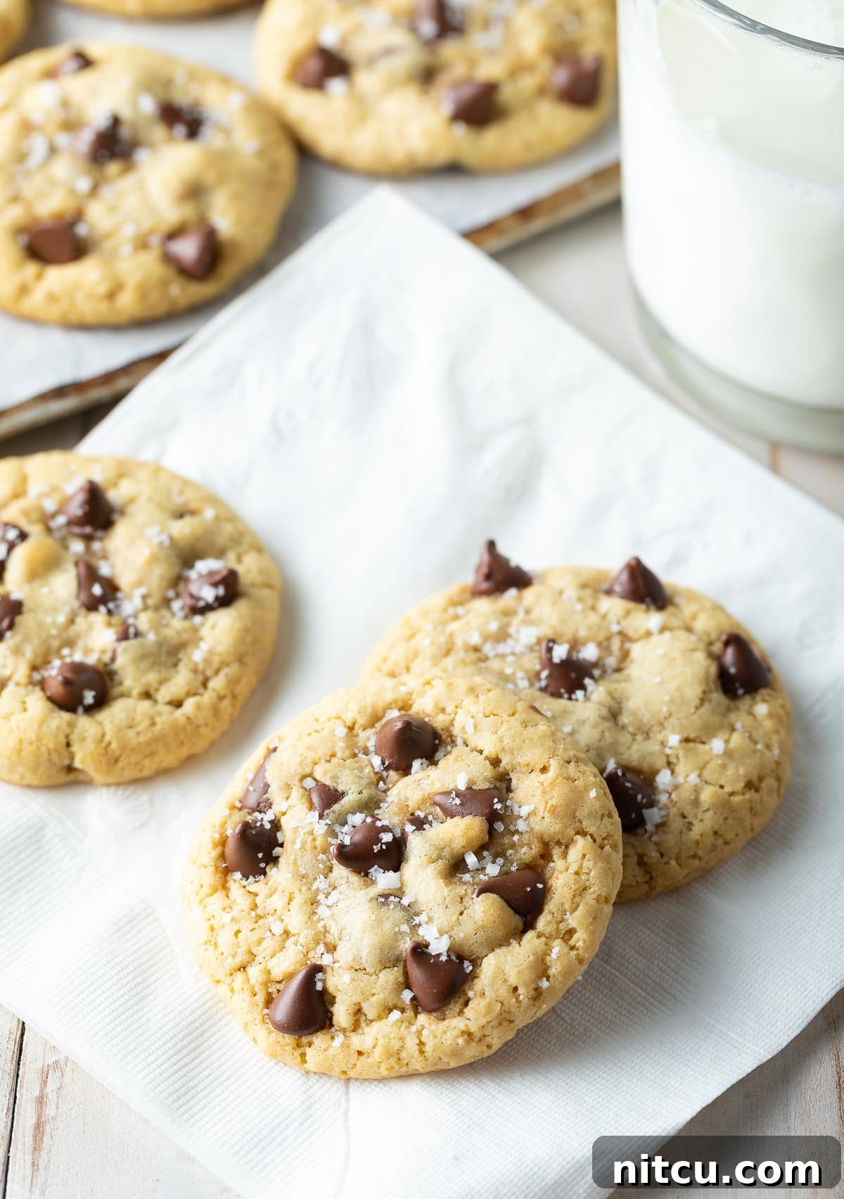Three perfectly baked salted chocolate chip cookies arranged on a pristine white napkin, showcasing their golden edges and melted chocolate.