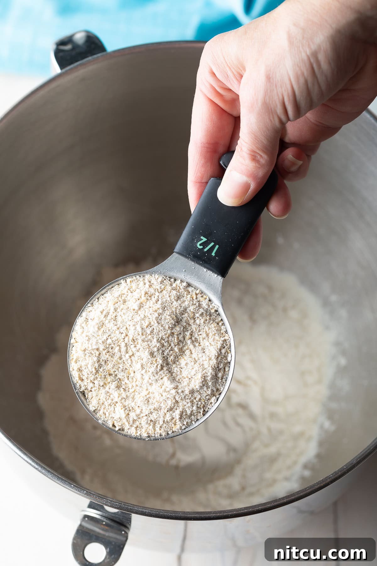 A hand using a measuring scoop to add oat flour to a metal mixing bowl, part of the dry ingredient combination.