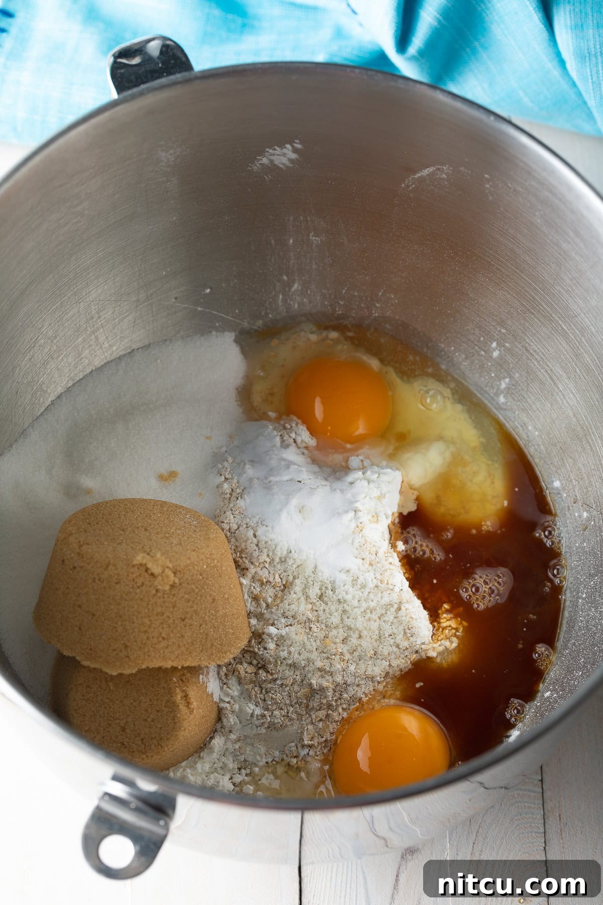 A metal mixing bowl filled with dry ingredients, brown sugar, white sugar, eggs, and vanilla extract, ready to be mixed into cookie dough.
