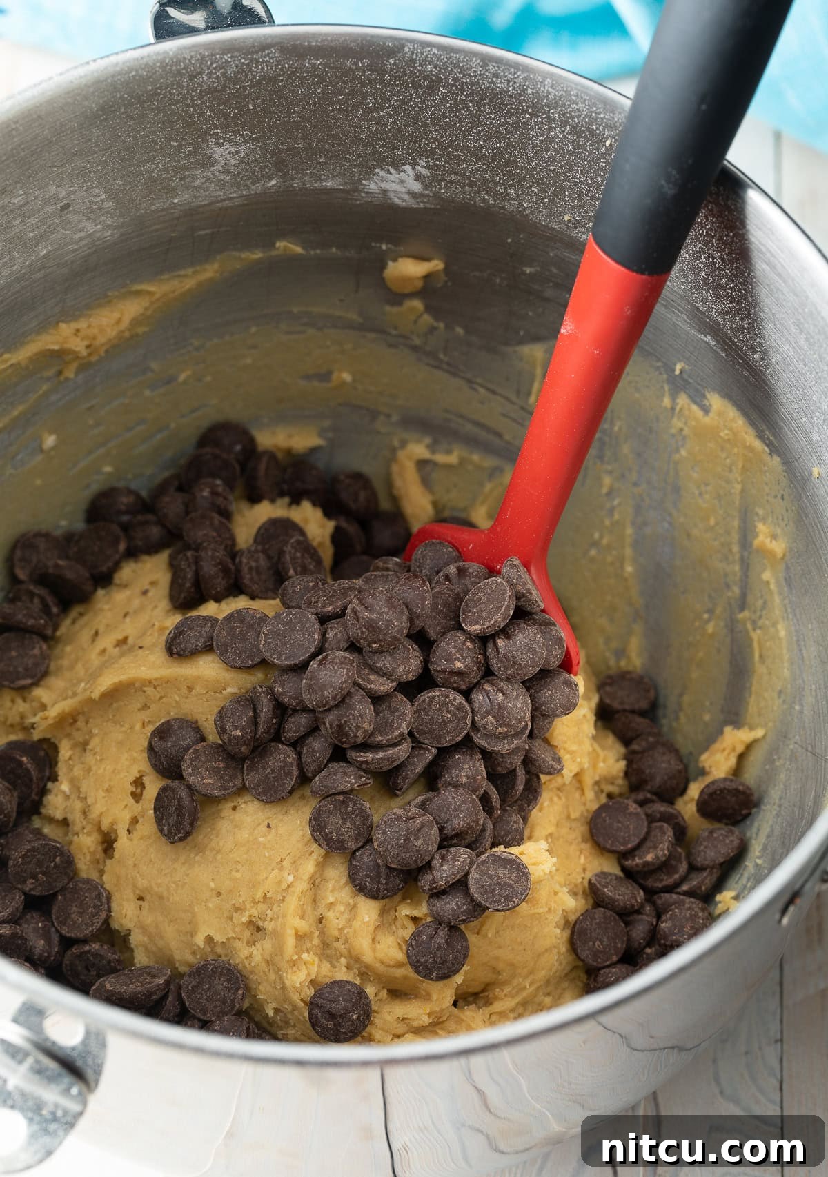 A red spatula stirring generous amounts of chocolate chips into a bowl of rich, thick cookie dough.