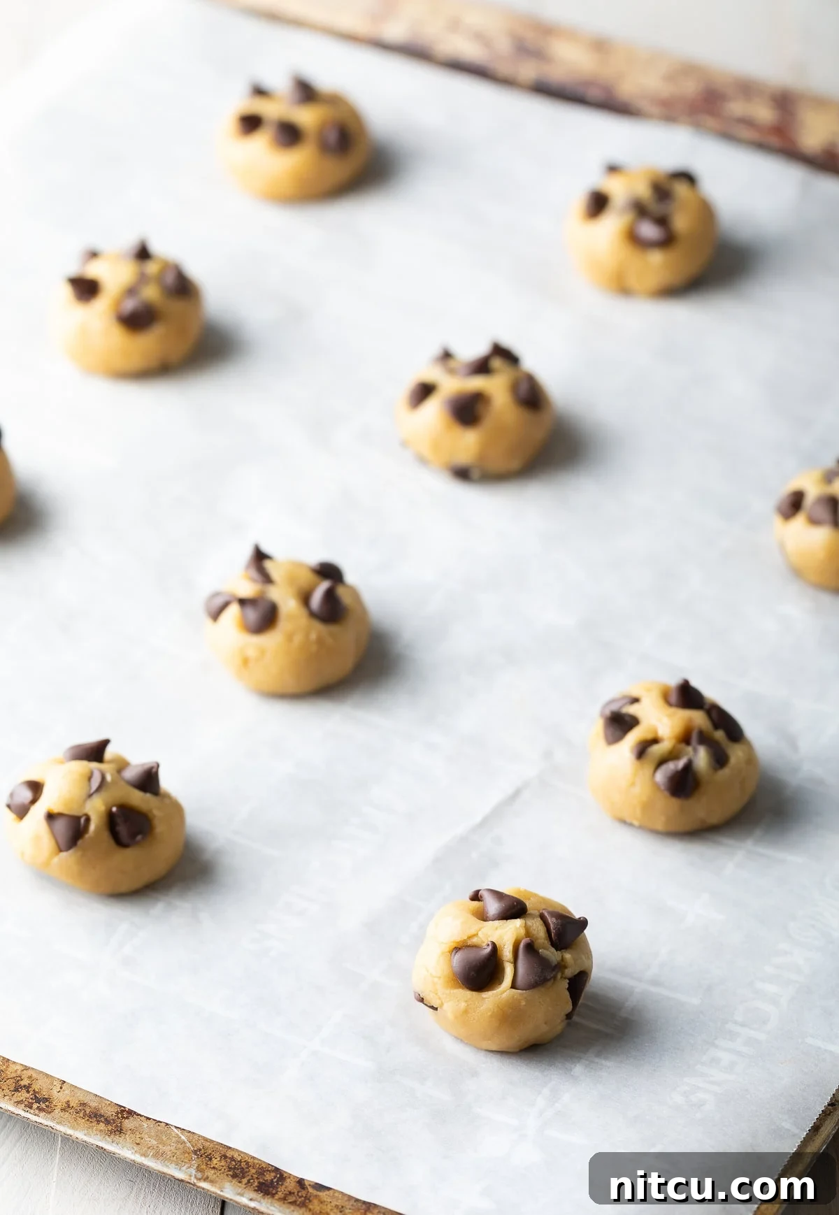 A dozen raw chocolate chip cookie dough balls, topped with extra chips, neatly arranged on a parchment-lined baking sheet, ready for the oven.