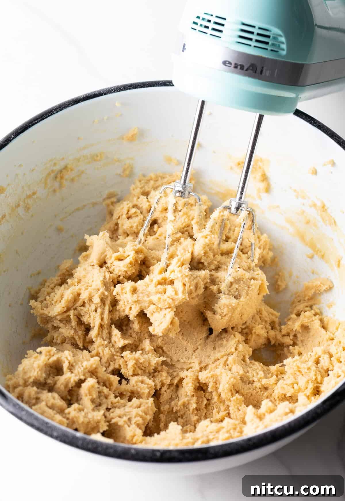 A close-up of Milky Way cookie dough in a white bowl being blended with an electric mixer, showcasing its smooth texture.