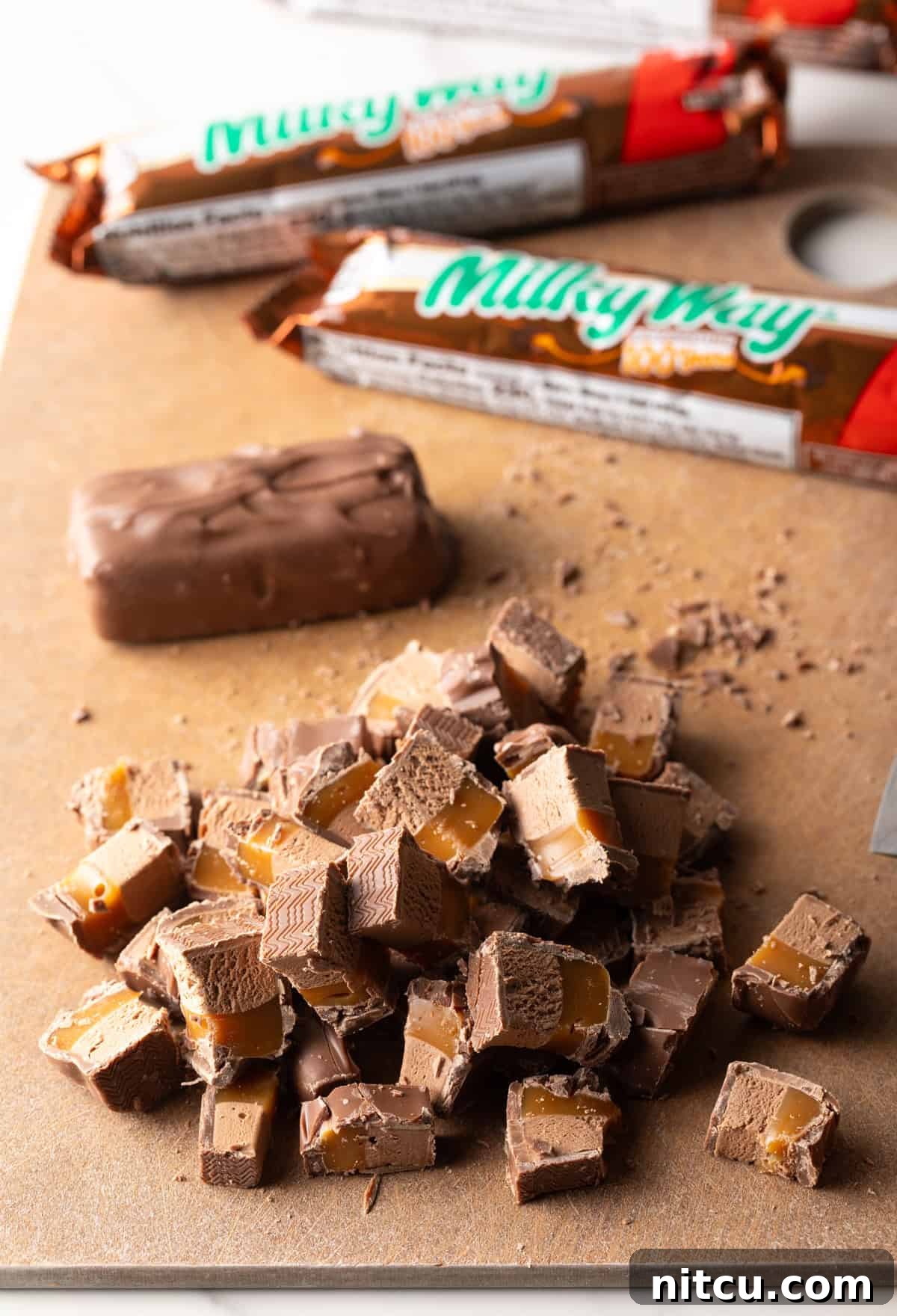 Pieces of chopped Milky Way bars on a wooden cutting board, ready to be added to cookie dough.