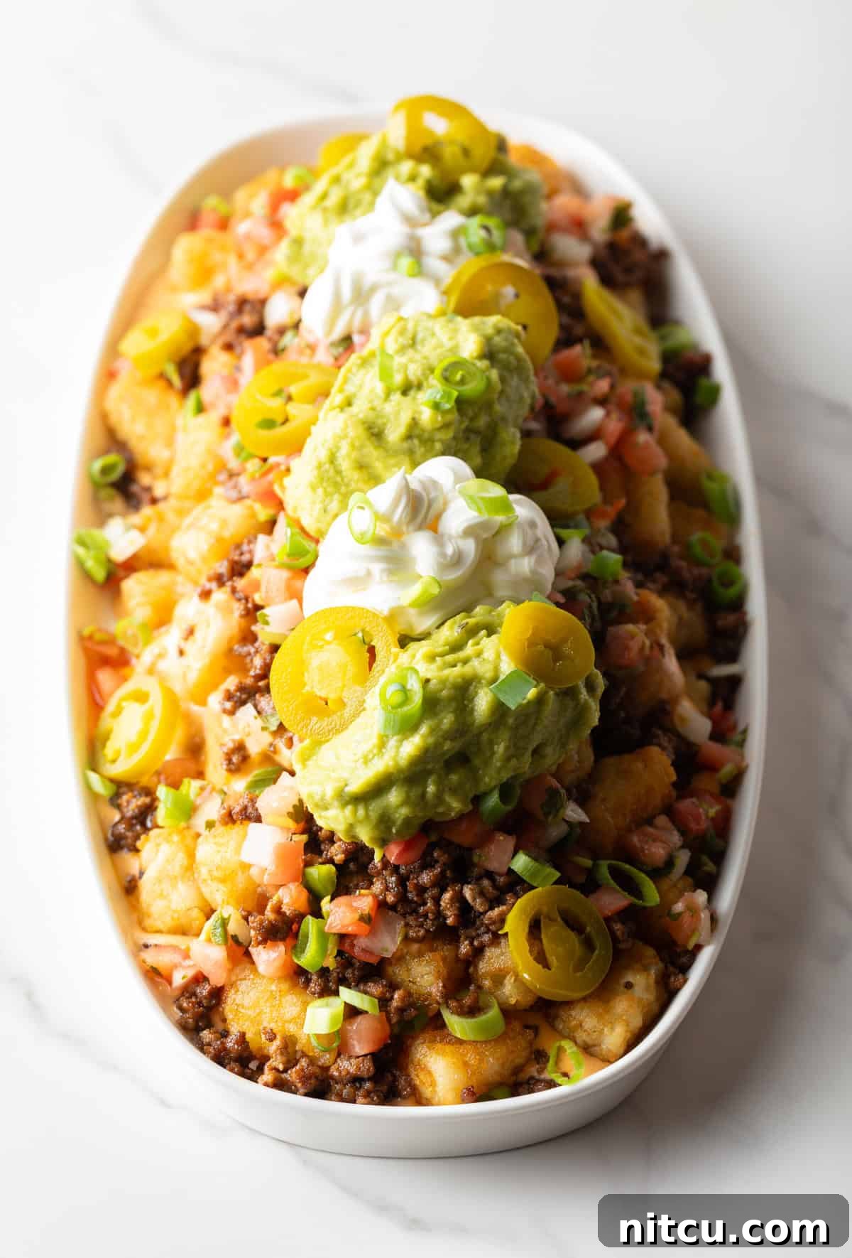 Top down view of large white oval serving dish loaded with tater tot nachos with ground beef, jalapenos, salsa, cheddar cheese, guacamole and sour cream.