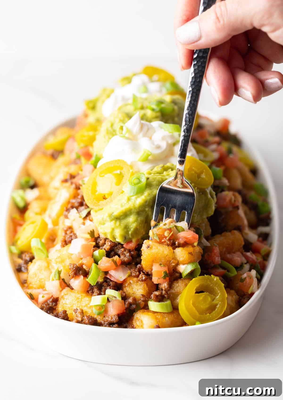 Hand with a fork spearing a bite of totchos from a large white oval serving dish.