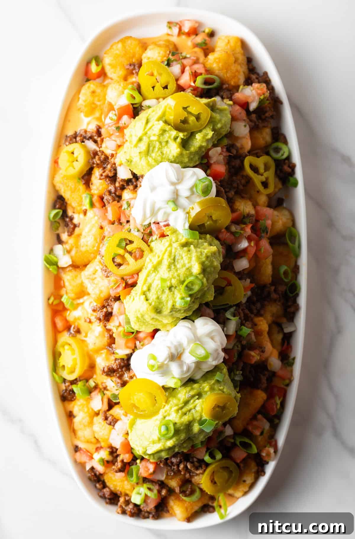 Top down view of large white oval serving dish loaded with tater tot nachos with ground beef, jalapenos, salsa, cheddar cheese, guacamole and sour cream.