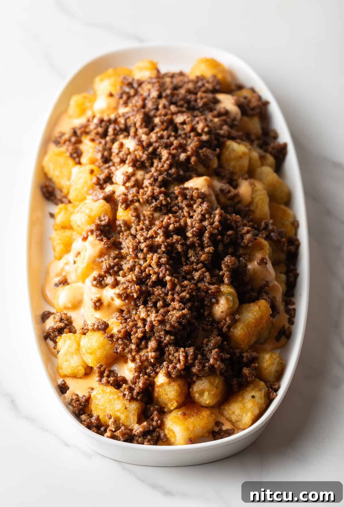 Tater tots in a large white oval bowl covered with ground beef.