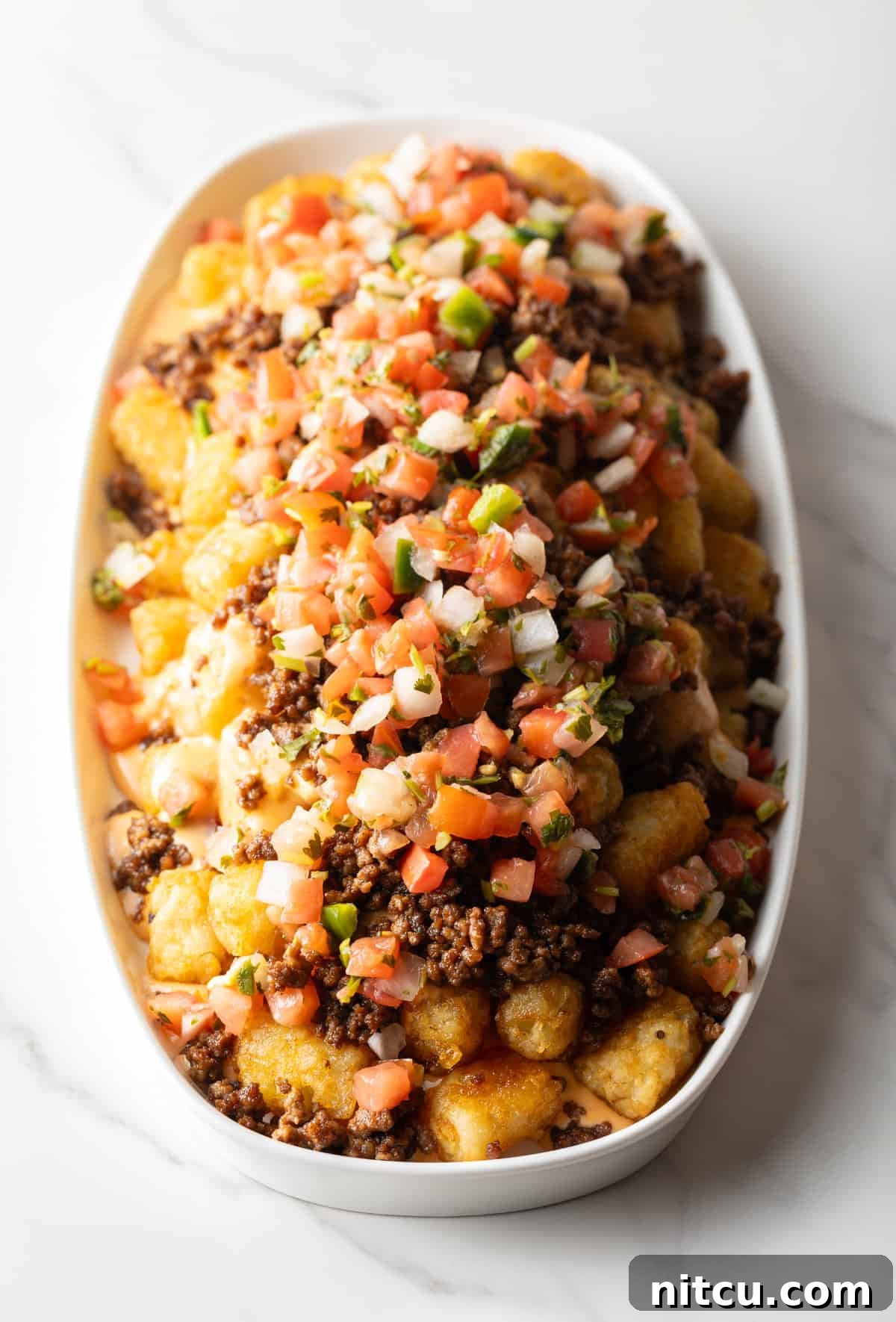 Tater tots in a large white oval bowl covered with ground beef and pico de gallo.