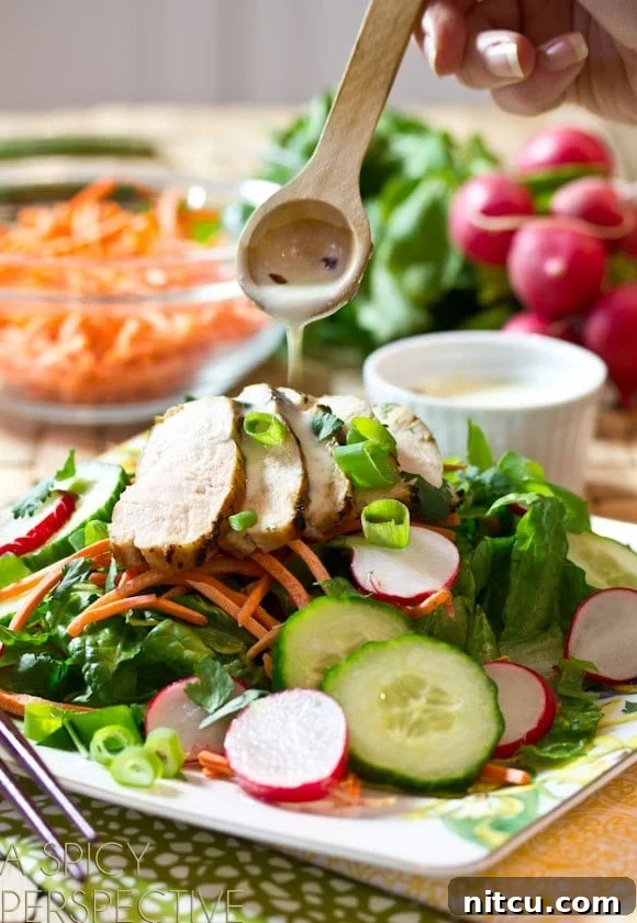 A close-up shot of a hand elegantly drizzling the creamy Vietnamese dressing over a vibrant plate of Banh Mi salad. The salad is loaded with crisp greens, bright pickled cucumbers and radishes, shredded carrots, and tender slices of grilled chicken breast, all awaiting the final touch of dressing.