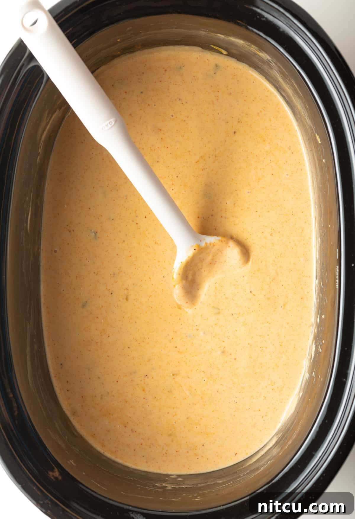 Black slow cooker filled with queso dip with a white spoon stirring it.