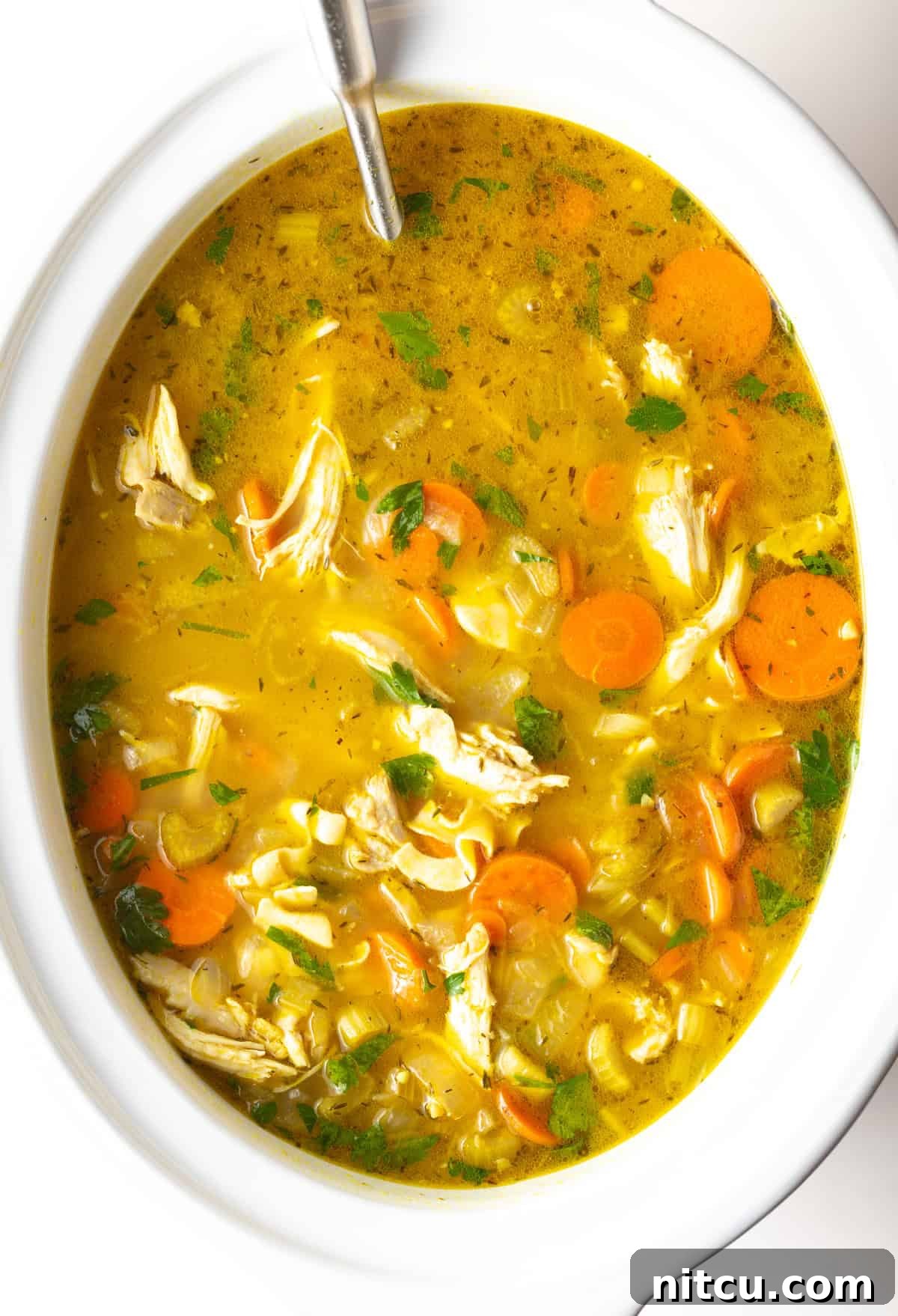 Chicken noodle soup in a white crockpot with a metal ladle.