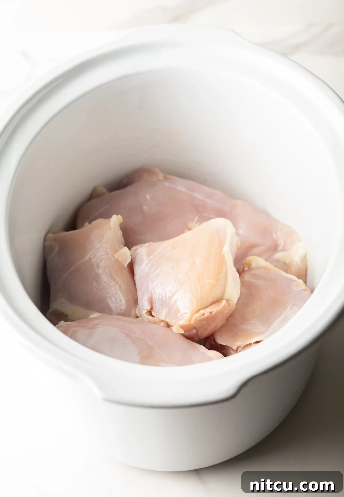 Raw chicken breasts are arranged in the base of a white crockpot.