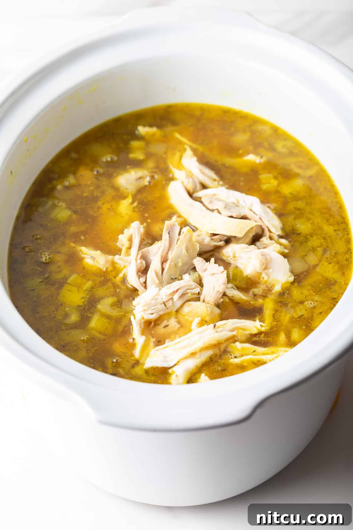 Shredded chicken being added to crockpot chicken noodle soup in a white slow cooker.