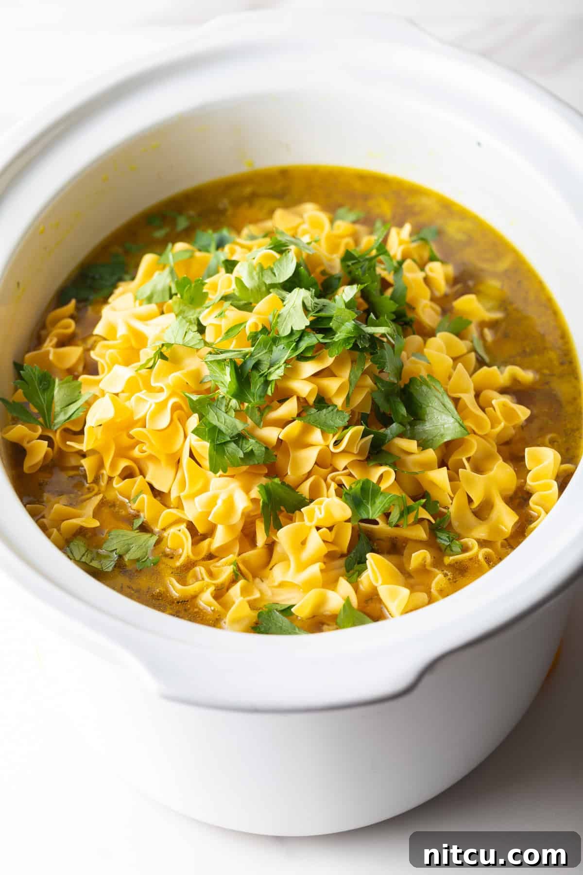 Uncooked egg noodles and parsley are being added to the broth mixture for slow cooker chicken noodle soup. 