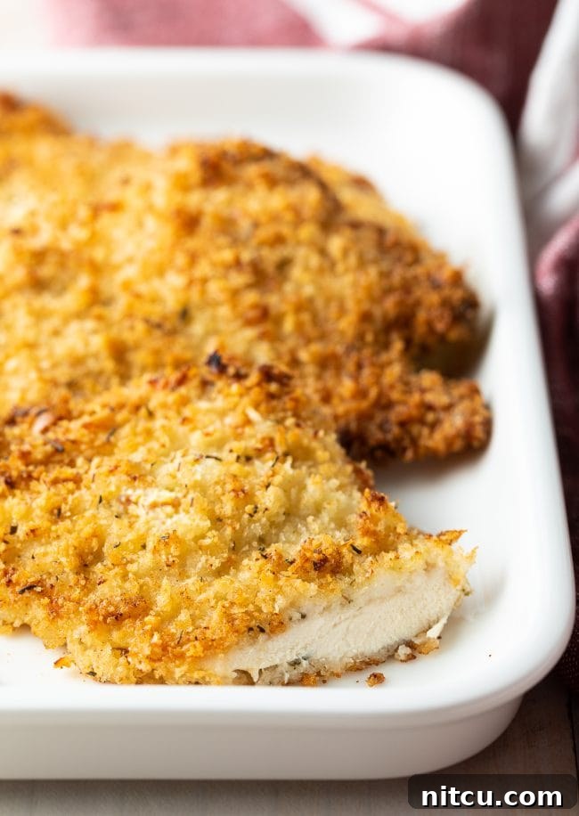 Baked chicken cutlet on a white platter with a slice removed, showing its juicy interior.