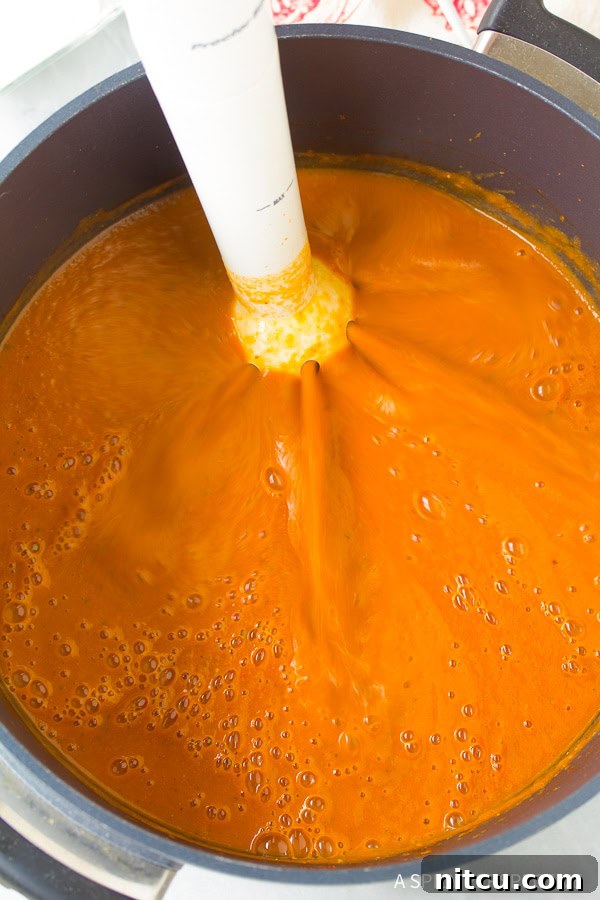 Healthy tomato basil bisque simmering in a pot with an immersion blender blending the ingredients to a smooth consistency.