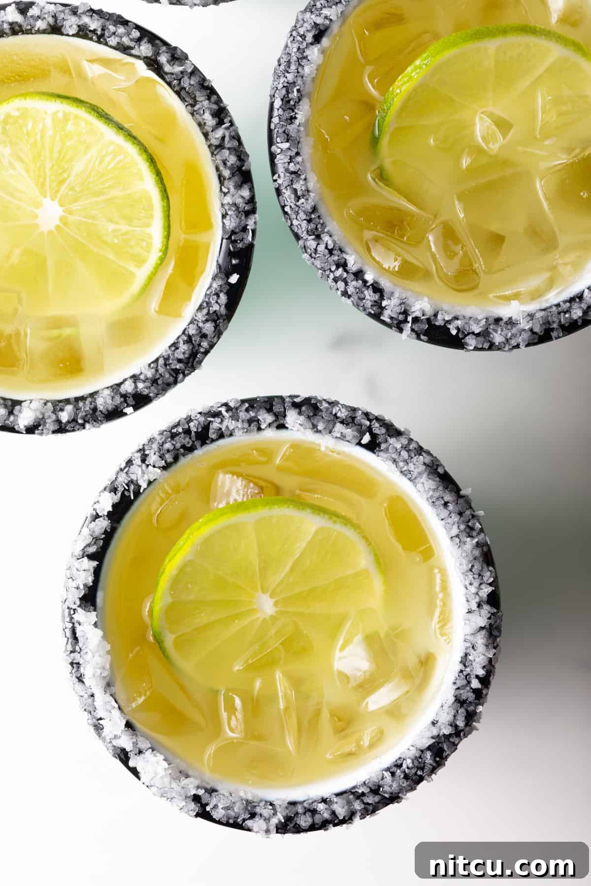 Top down view of 3 white and green-striped cups with margaritas on the rock, with salt around the rim and a slice of lime in the drink.