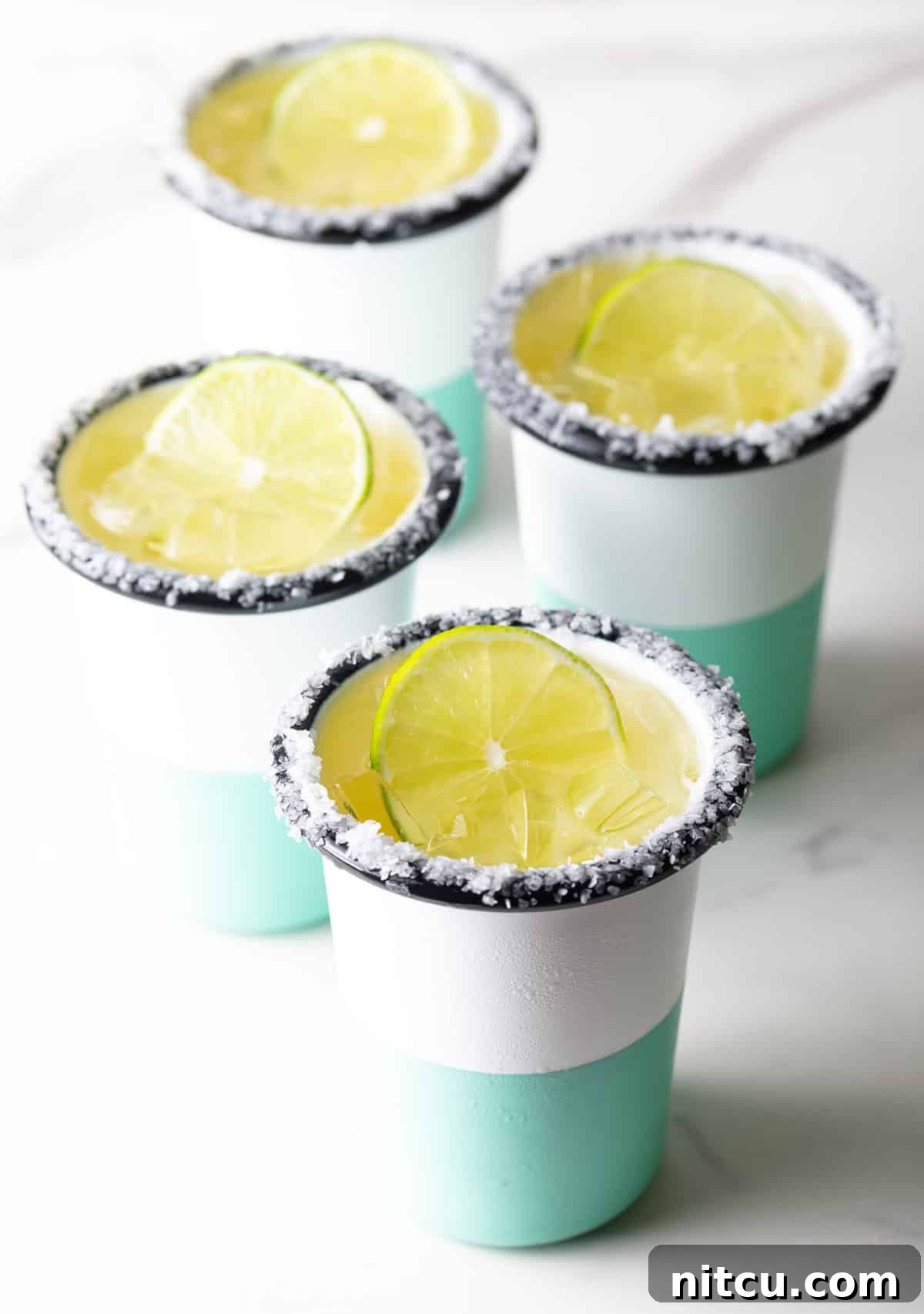 4 white and green-striped cups with margaritas on the rock, with salt around the rim and a slice of lime in the drink.