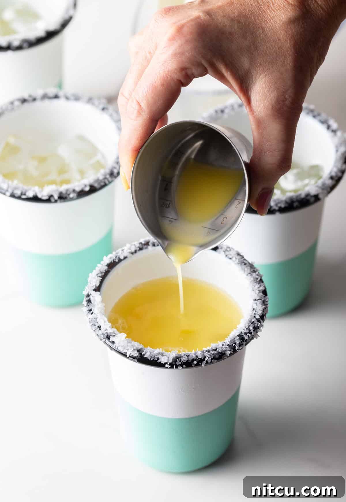 Hand pouring juice from a metal shot glass into a cup with a salted rim.