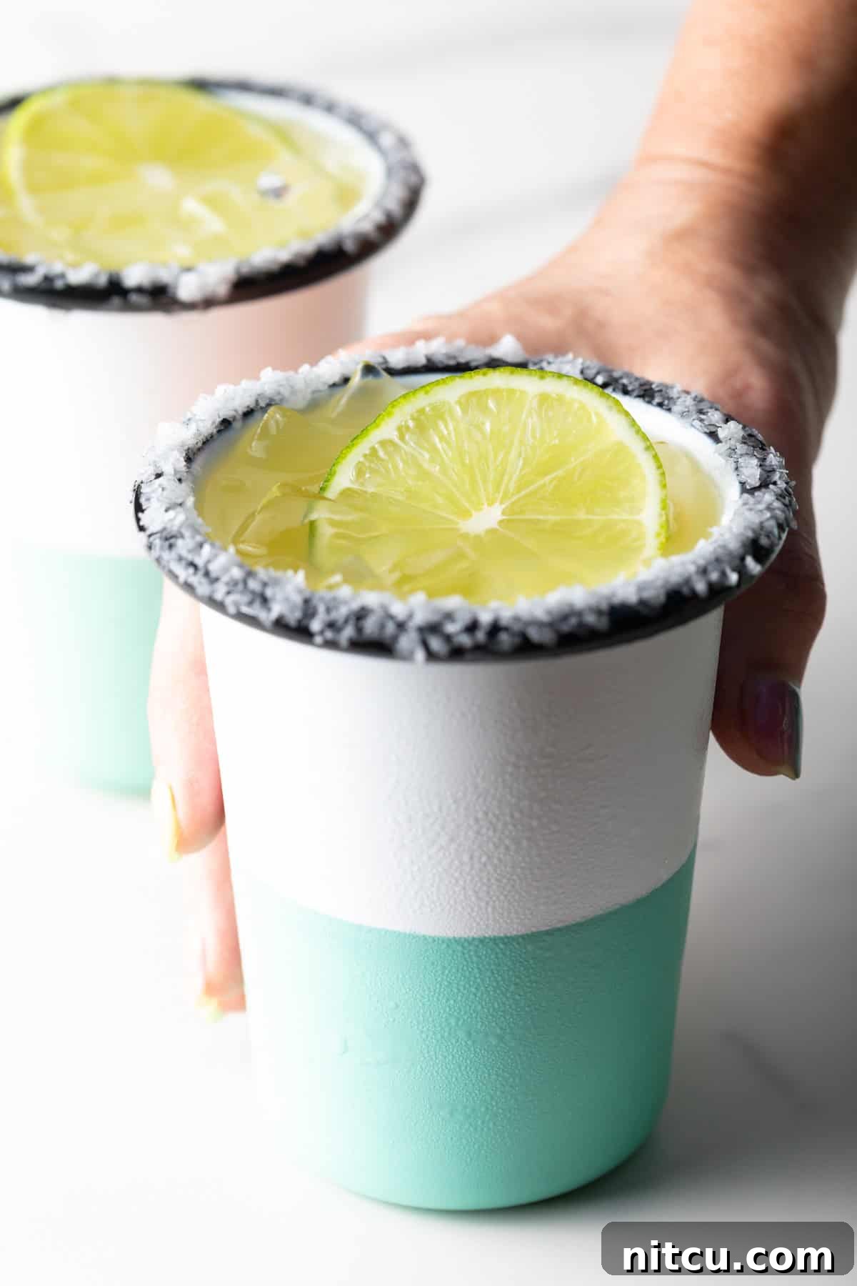 Hand grabbing a white and green-striped cup with salt around the rim and slice of lime in the drink.